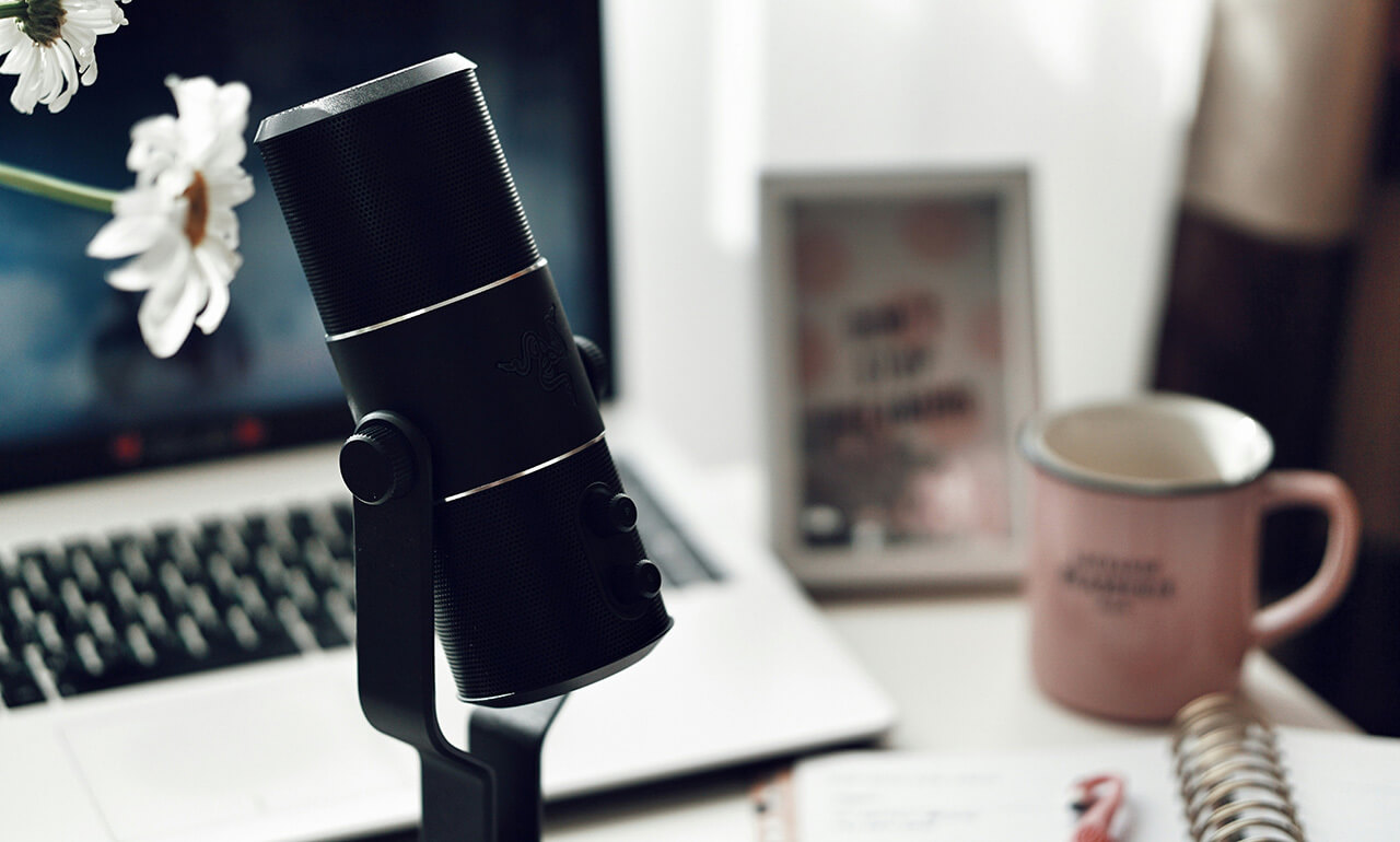 Ein Schreibtisch, auf dem im Vordergrund ein Mikrofon zu sehen ist. Im Hintergrund sind ein ein Notebook, ein Kaffeebecher und ein Notizblock mit Stift zu sehen.